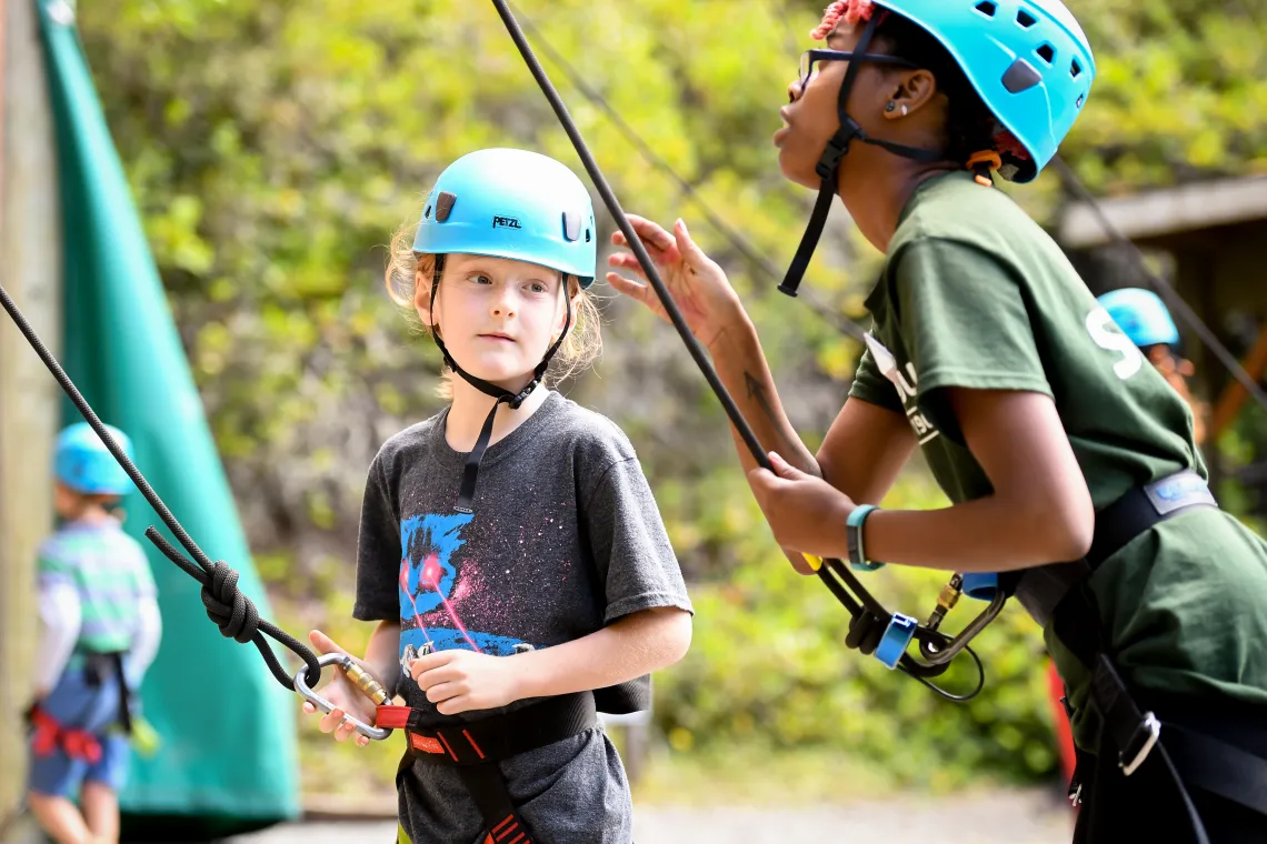 ) A child wearing a blue helmet listens to an instructor, also wearing a helmet, at an outdoor climbing activity. Both are in harnesses, discussing climbing techniques. Trees and climbing equipment are visible in the background.