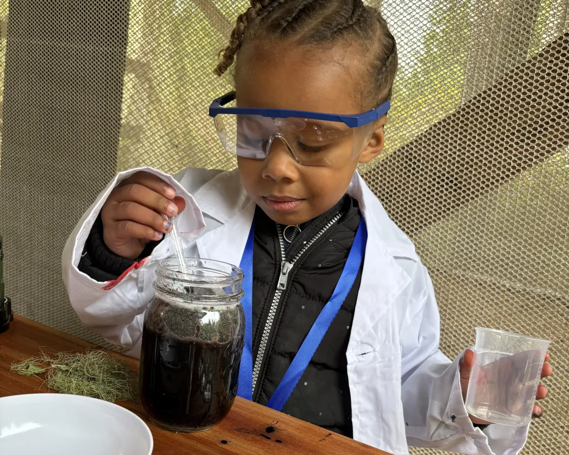 A young child wearing safety goggles and a lab coat observes a jar filled with dark liquid, pouring water from a cup into it. The child is at a wooden table with spoons and a white bowl nearby.