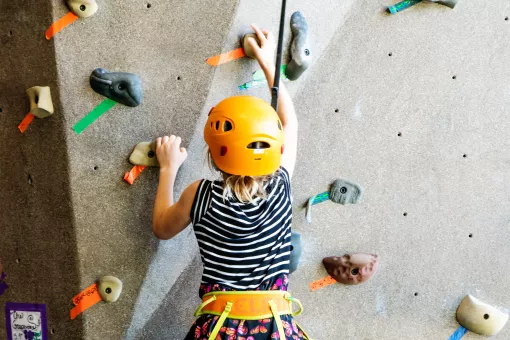 Kids is focusing climbing up a rock wall in an orange harness and helmet.