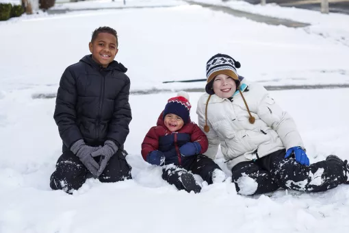 Three kids sit in the snow, two middle school age and one toddler.