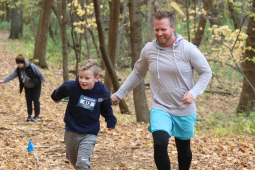 A father in son race up a trail, smiling as they move across fallen leaves on a cold day.