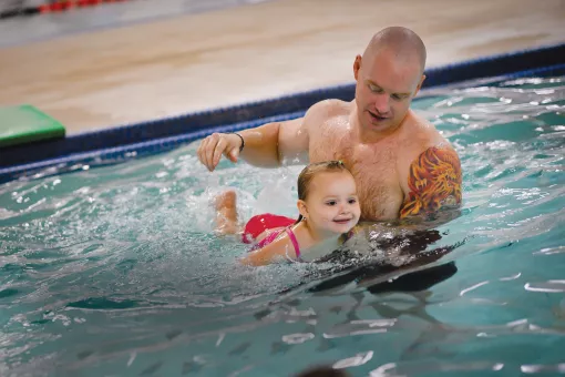 A father holds his young daughter up in the water as they leans on a kickboard and creates splashes behind her. 