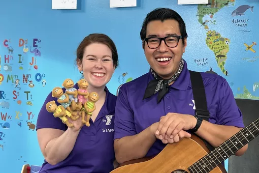 YMCA360 instructors Lauren and Gabriel smile toward the camera in matching purple collared shirts. Lauren holds up 4 moneky finger puppets in pastel dresses. Gabriel wears a handkerchief and rests his hands on an acoustic guitar.