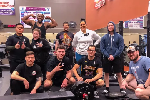 A group of adults huddle together and smile while inside the Tacoma Center YMCA weight room. 
