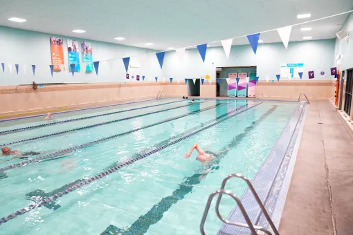 Indoor lap pool at the Tacoma Center YMCA with multiple swim lanes, lane dividers, and triangular flags overhead. Several swimmers use the lanes, with pool ladders and tiled deck visible along the edge.