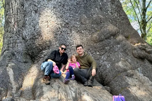 Sarah, her daughter Nora, and her husband sit together at the base of a large tree during a family outing.