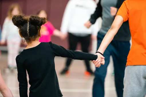 A child holds hands with an adult while standing in a gymnasium, with several other people blurred in the background participating in a group activity.