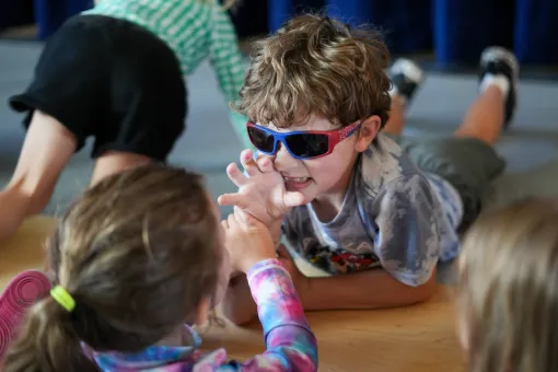 Children participating in an interactive educational activity indoors, with one child wearing colorful glasses while engaging with peers on a classroom floor during a youth learning program.