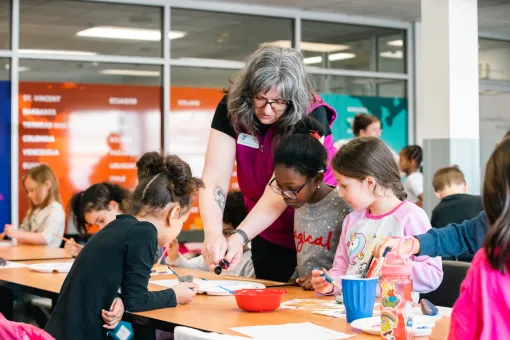 An instructor leans over a table to assist several children with an arts and crafts project at a YMCA classroom. Participants are seated around the table drawing and painting, with art supplies, cups, and paper spread across the workspace. Large windows and colorful wall displays are visible in the background.