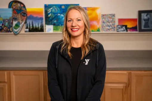Michelle Wells, Arts Director, standing indoors in front of a display of colorful artwork and paintings. Wearing a dark YMCA-branded jacket over a black shirt, with light wood cabinetry visible below the art display.