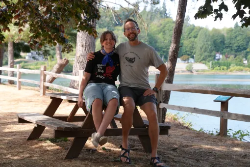 Alt text:  Aaron and Ryan sit together on a wooden picnic table at Camp Seymour beside the waterfront. Trees shade the area, with a wooden fence, calm water, and greenery visible in the background on a sunny summer day.