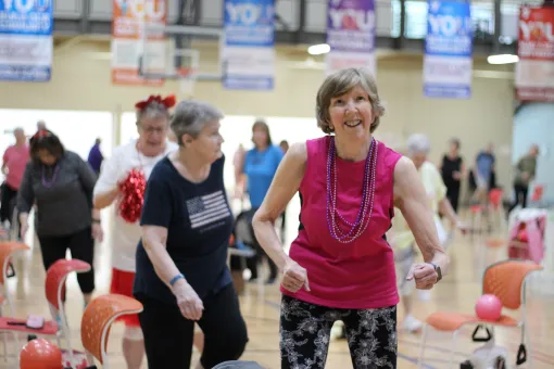 Adult participants take part in a group fitness class at a YMCA gymnasium. One participant in a bright pink sleeveless top exercises near the front of the group, with chairs arranged throughout the room for support. YMCA banners hang in the background above the activity area.