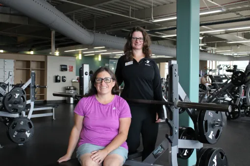 Julia poses with a YMCA staff member in the fitness center at the Gordon Family YMCA. Julia sits on a weight bench in front of a barbell while the staff member stands beside the bench. Strength training equipment and exercise machines are visible in the background.