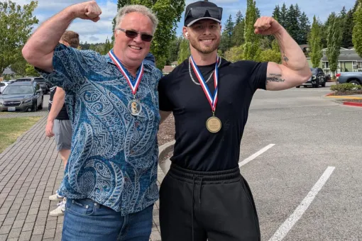 Two medal-winning participants flex their arms after the Haselwood Family YMCA Strongman Competition, standing outdoors in a parking lot following the community strength event in Silverdale, Washington.