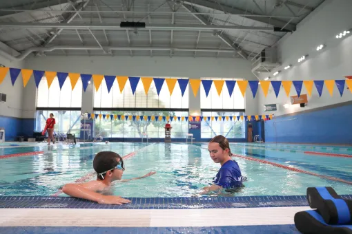 Young swimmer practicing skills during a private swim lesson with an instructor in an indoor YMCA pool. The pair are positioned near the pool edge, with lap lanes, blue and gold pennant flags, and large windows visible in the background.