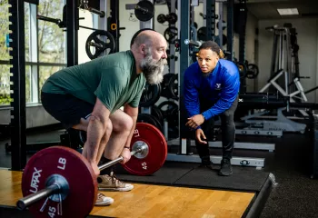 A man prepares to perform a deadlift with a Y staff member nearby in blue watching alongside for his form.
