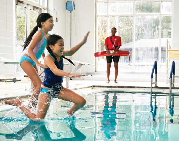 Two young girls jump off the edge of the pool and into the water while a lifeguard watches. 