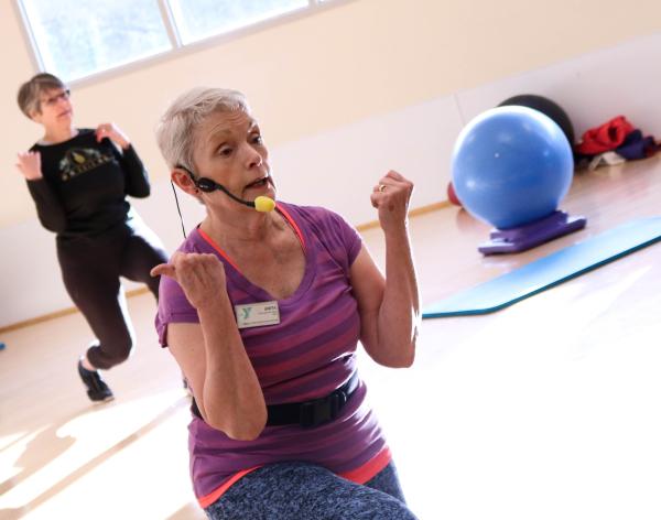An active older instructor teaches core movement to a group of participants in a sun filled studio. 