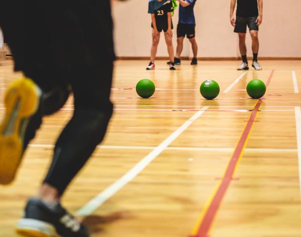 A row of green dodgeballs line up in the center of a basketball court as a player runs up to grab one. 