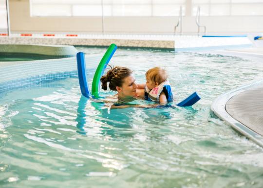 Woman holds a baby in the water. Baby is wearing a lifejacket and there are two pool noodles around them.