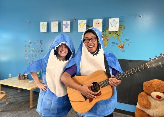 Instructors Lauren and Gabriel are in shark costumes and smiling as Gabriel holds an acoustic guitar.
