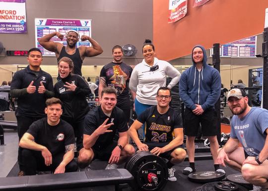A group of adults huddle together and smile while inside the Tacoma Center YMCA weight room. 