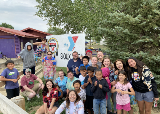 Summer of Service participants pose by a sign created in partnership with the YMCA of the Sioux Tribe.