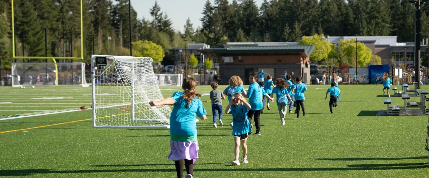 Children in blue YMCA shirts run and play on the new synthetic turf fields at the Gig Harbor Sports Fields following the ribbon‑cutting celebration, with soccer goals and trees in the background.