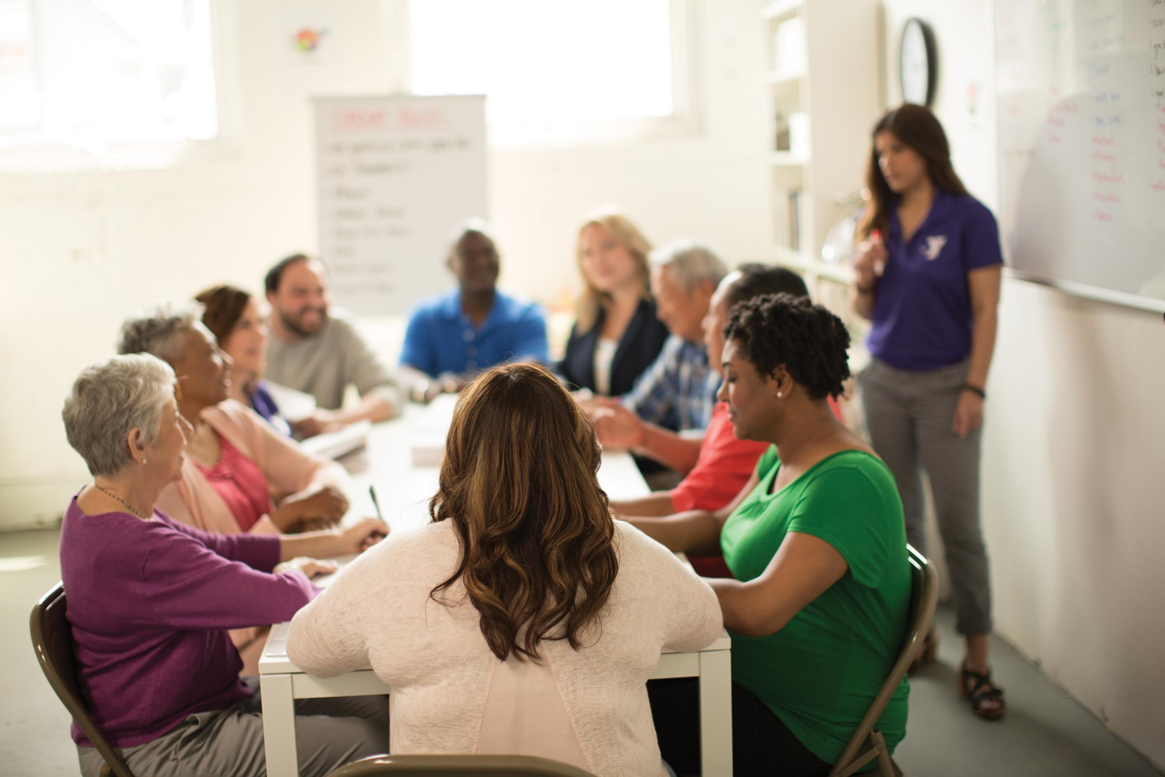 Diverse group of people sitting at a long table in the sunlight talking. Alongside them is a person near a white board with a YMCA polo shirt on.