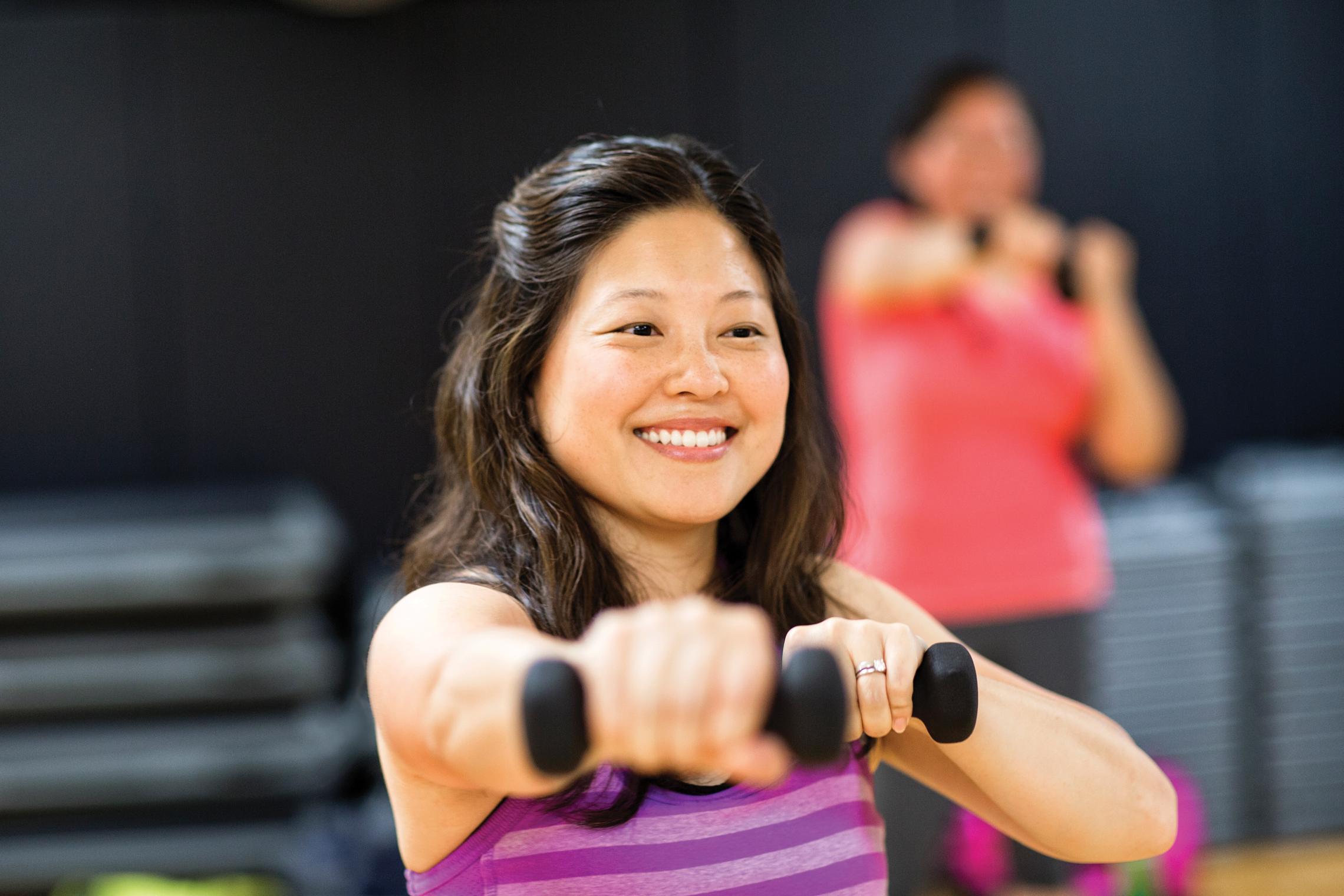 A young woman smiles in a purple striped tank top and lifts hand weights in a group exercise class. 