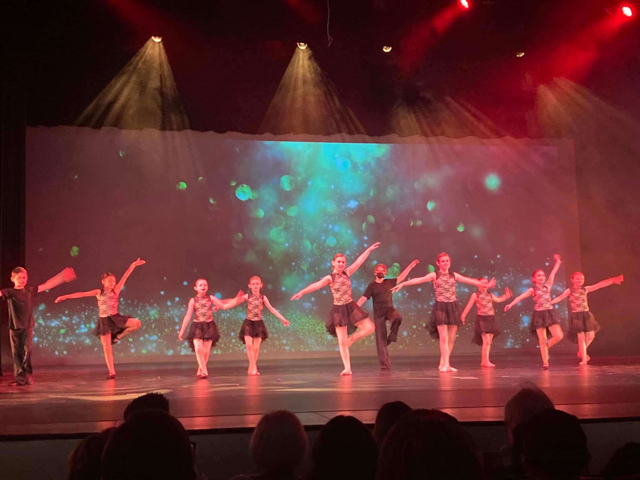 A group of young dancers performs on a theater stage during a recital. The performers are arranged in a line with arms extended in dance poses under red stage lighting. A colorful backdrop with blue and green light effects fills the background while audience members are visible in silhouette at the bottom of the image.