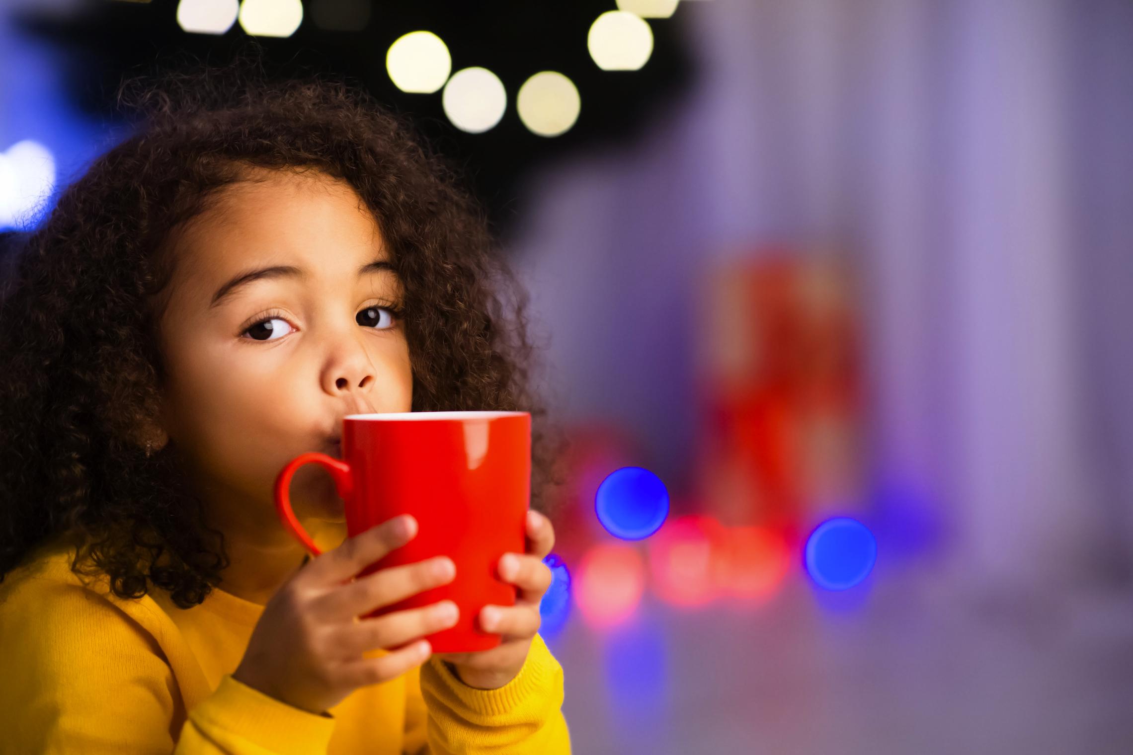 Young child sips hot cocoa in front of holiday lights.