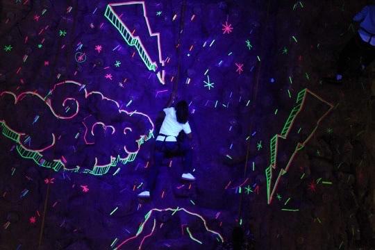 A climbing wall is illuminated by neon drawings as a Y member scales upward in the dark. 