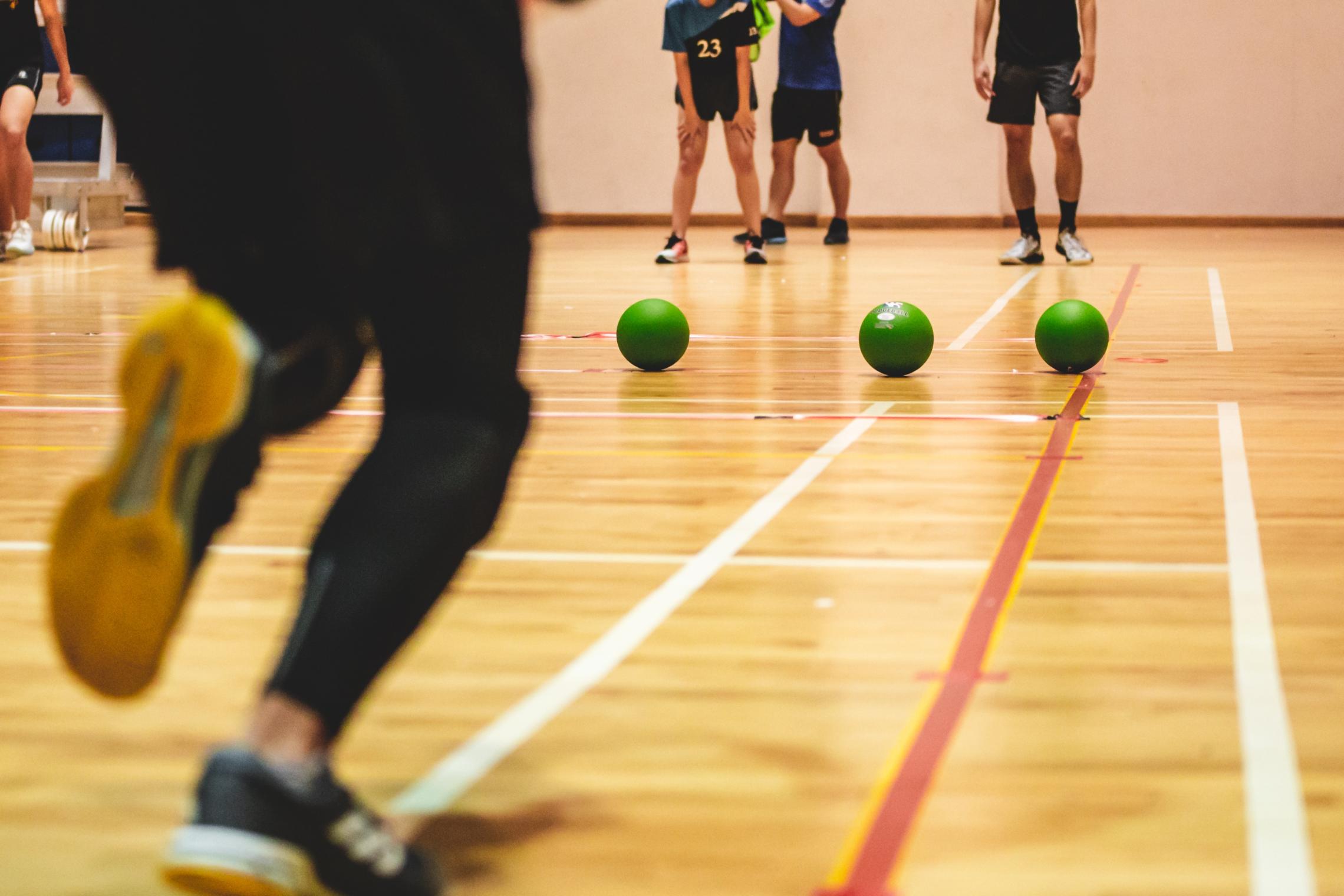 A row of green dodgeballs line up in the center of a basketball court as a player runs up to grab one.