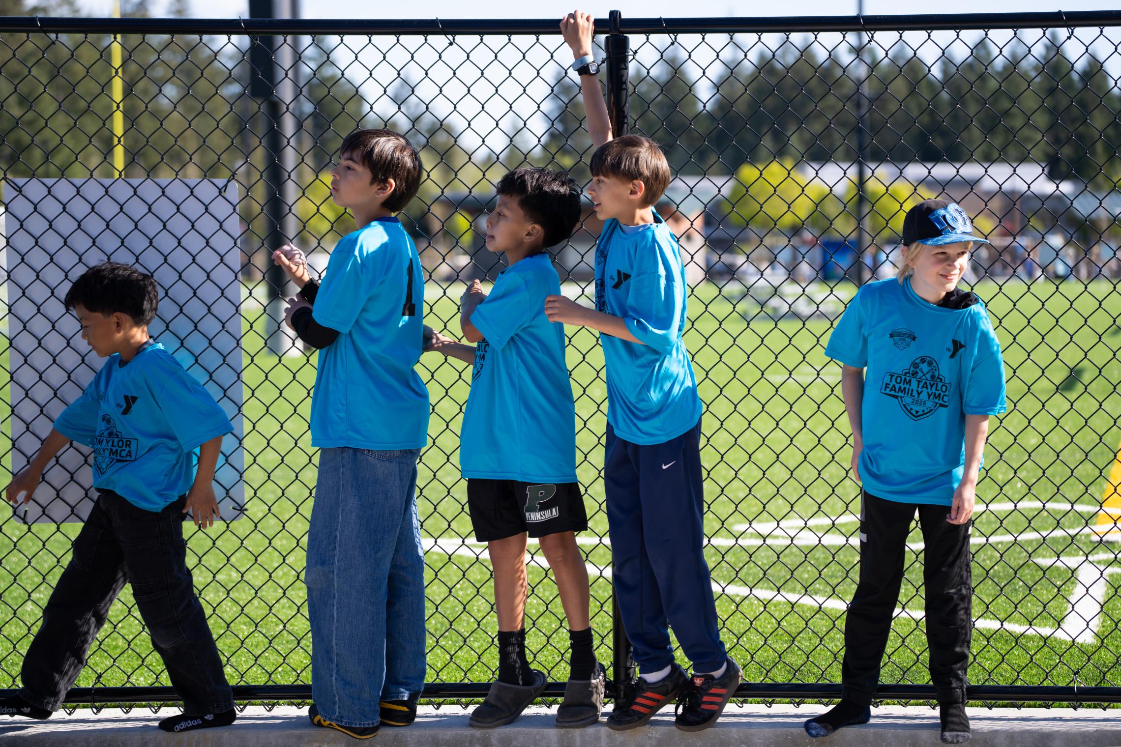 Children in blue YMCA shirts stand along a chain‑link fence overlooking the Gig Harbor Sports Fields during the opening celebration, with green turf fields visible in the background.