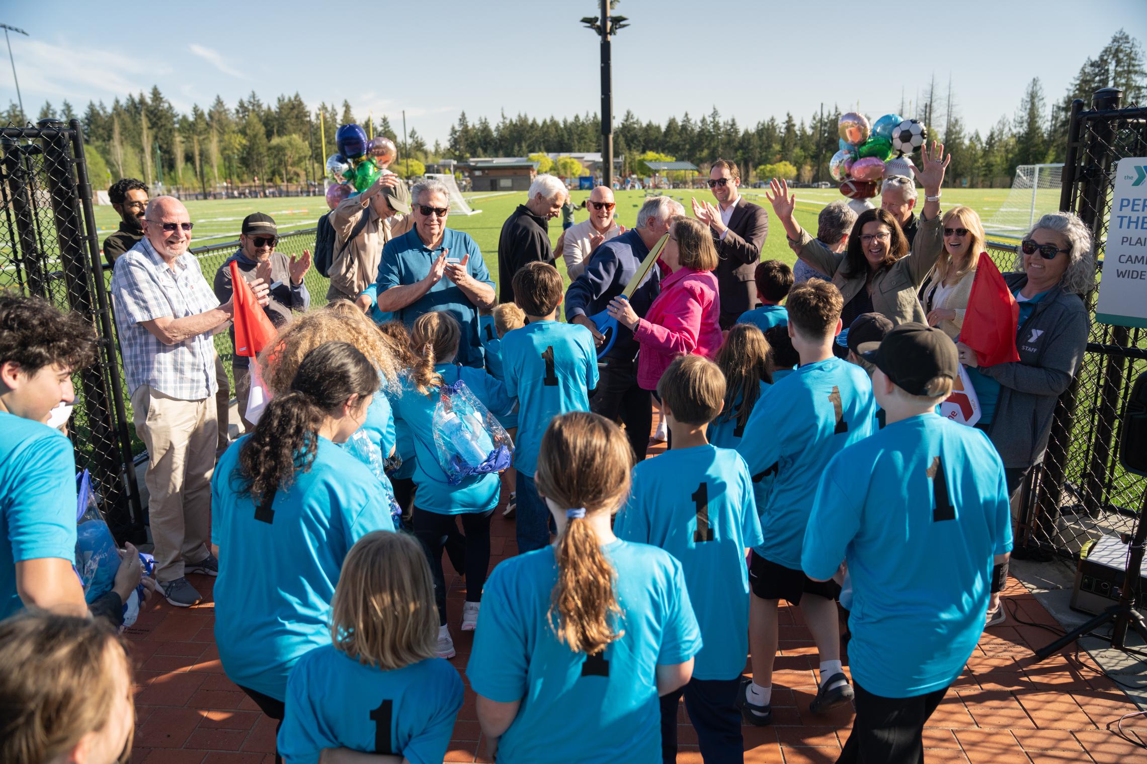 Community members and children wearing blue YMCA shirts gather and applaud during the ribbon‑cutting celebration at the Gig Harbor Sports Fields, with balloons and turf fields visible behind them.