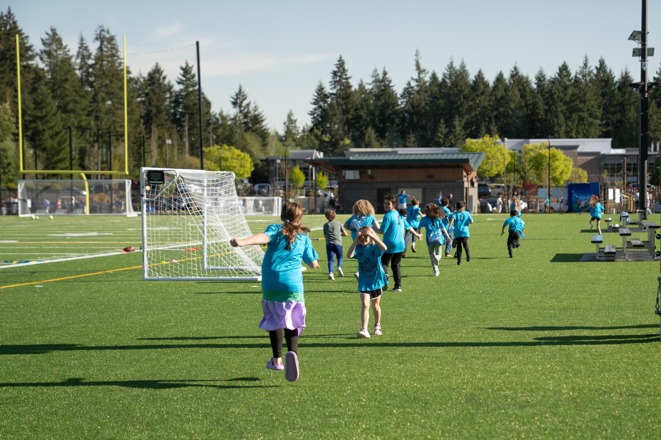 Children in blue YMCA shirts run and play on the new synthetic turf fields at the Gig Harbor Sports Fields following the ribbon‑cutting celebration, with soccer goals and trees in the background.