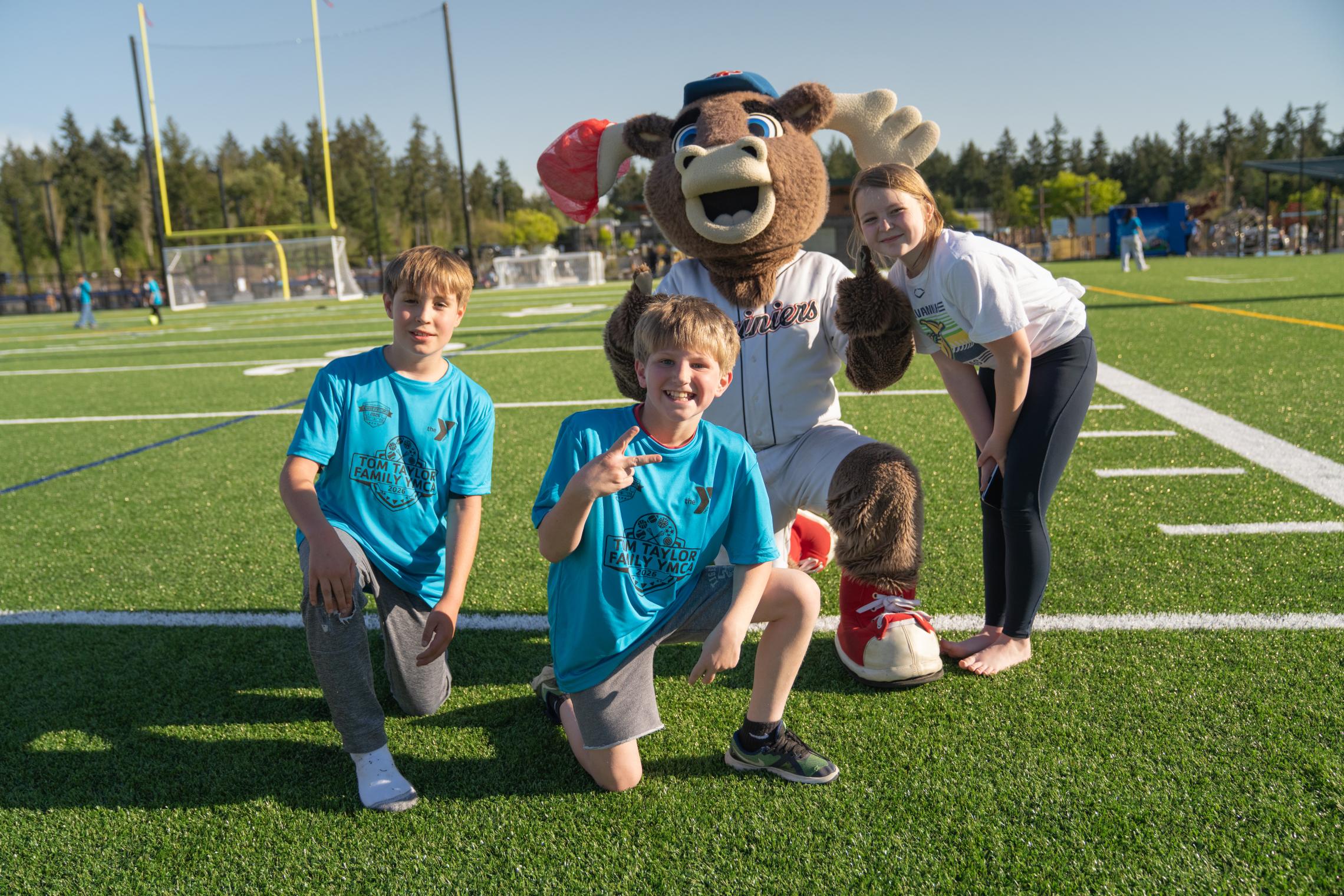 Children wearing blue YMCA shirts pose on the turf field with Rhubarb, the Tacoma Rainiers mascot, during the Gig Harbor Sports Fields ribbon‑cutting celebration.