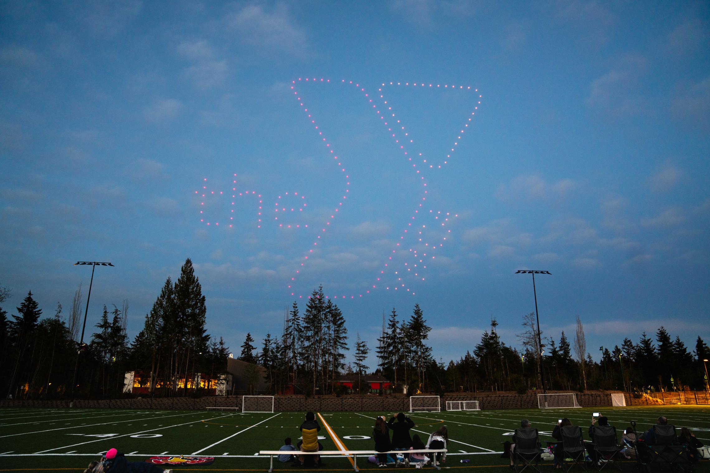 A drone light show displays the YMCA logo and the words “the Y” in the evening sky above the Gig Harbor Sports Fields during the ribbon‑cutting celebration, with spectators seated along the field below.