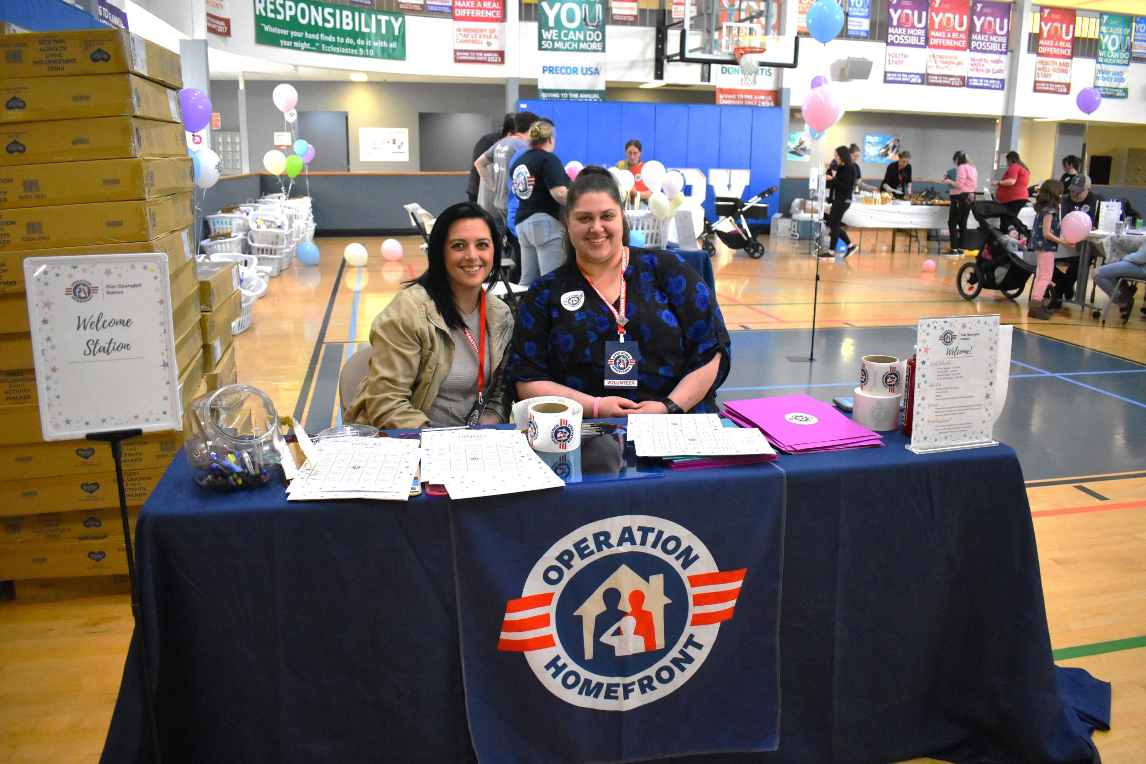 Operation Homefront welcome table at a gym during a community baby shower event, with two staff members seated behind a table covered in an Operation Homefront banner, sign-in materials, and boxes of supplies visible nearby.