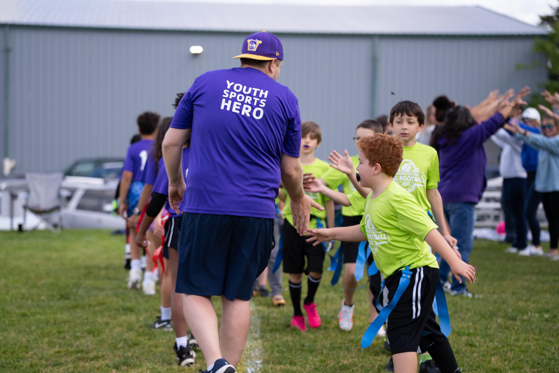 A volunteer in a purple shirt with the words “YOUTH SPORTS HERO” high-fives children lined up on a grassy field during a youth flag football event. Participants wear brightly colored jerseys and flag belts, while additional volunteers and spectators stand in the background.