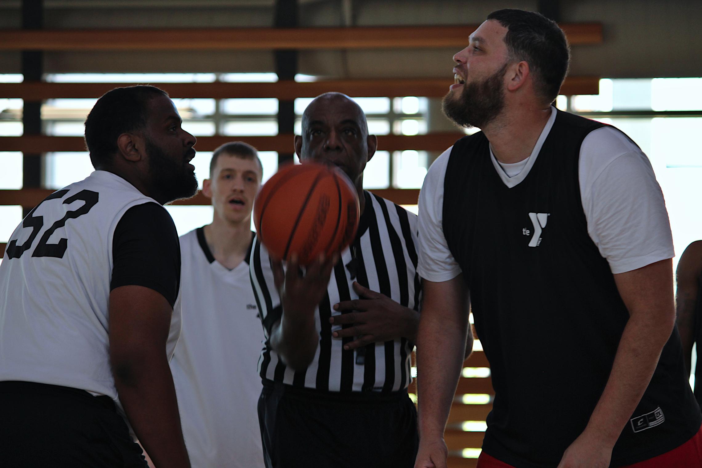 A referee tosses a basketball for the opening tip-off as two players face each other on an indoor basketball court at the University YMCA Student Center.