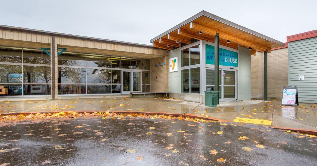 Alt text:  Exterior entrance of the Bremerton Family YMCA on a rainy day, featuring large glass windows, YMCA branding, a covered entryway with wood accents, and wet pavement scattered with autumn leaves.