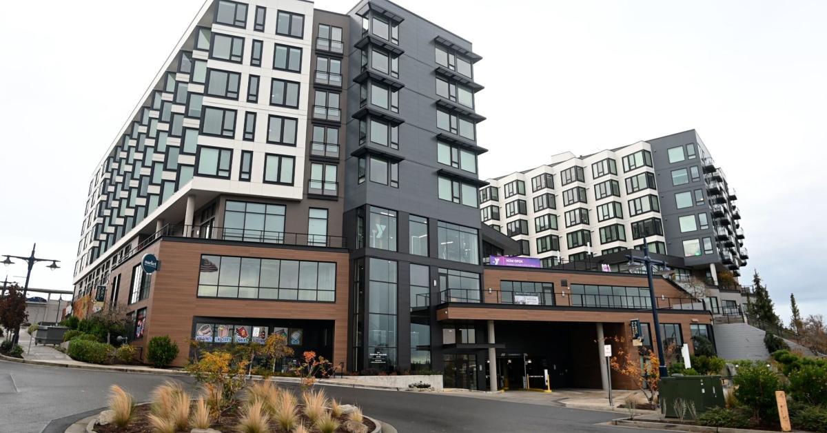 Exterior view of the Marina Square Express YMCA in downtown Bremerton, located on the ground floor of the modern Marina Square mixed-use development. The image shows multi-story residential buildings above and around the YMCA space, with large windows, street-level landscaping, and nearby pedestrian walkways.