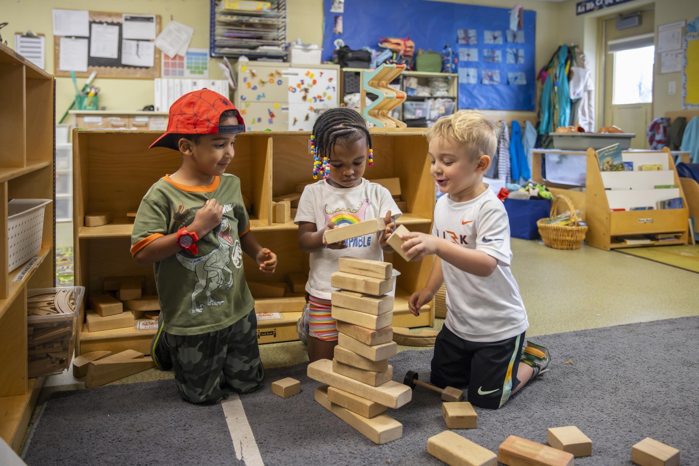 Children play with wooden building blocks in an early learning center classroom. The image shows a colorful learning environment with shelves, books, educational materials, and activity areas designed to support early childhood development and hands-on play.
