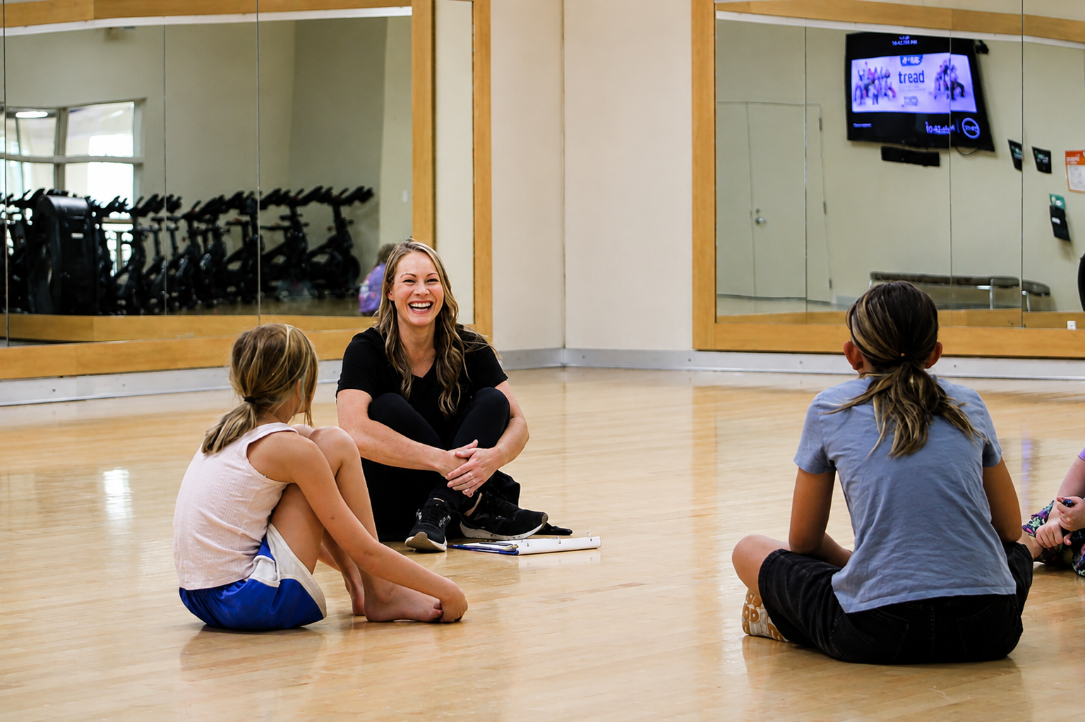 Michelle Wells, Arts Director, seated on a gymnasium studio floor leading a small group discussion with youth participants. The group sits in a circle in a fitness studio with mirrored walls and exercise equipment visible in the background.