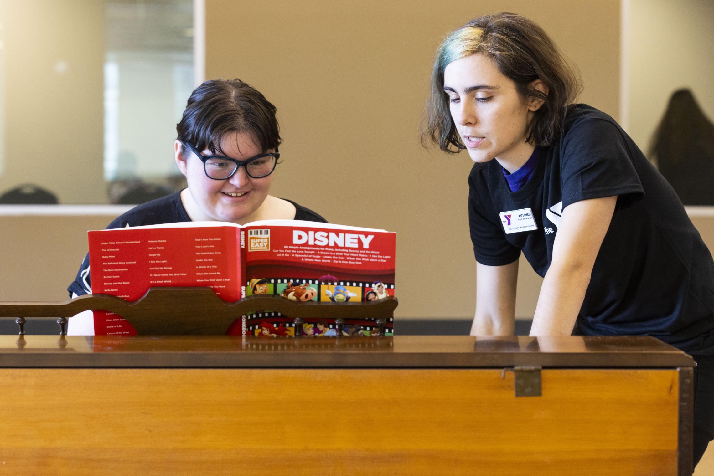 Two participants stand at a piano in a YMCA arts classroom, reviewing a Disney music book on a stand. One participant leans over the piano while the other looks at the sheet music, with a YMCA staff name badge visible and a neutral classroom setting in the background.