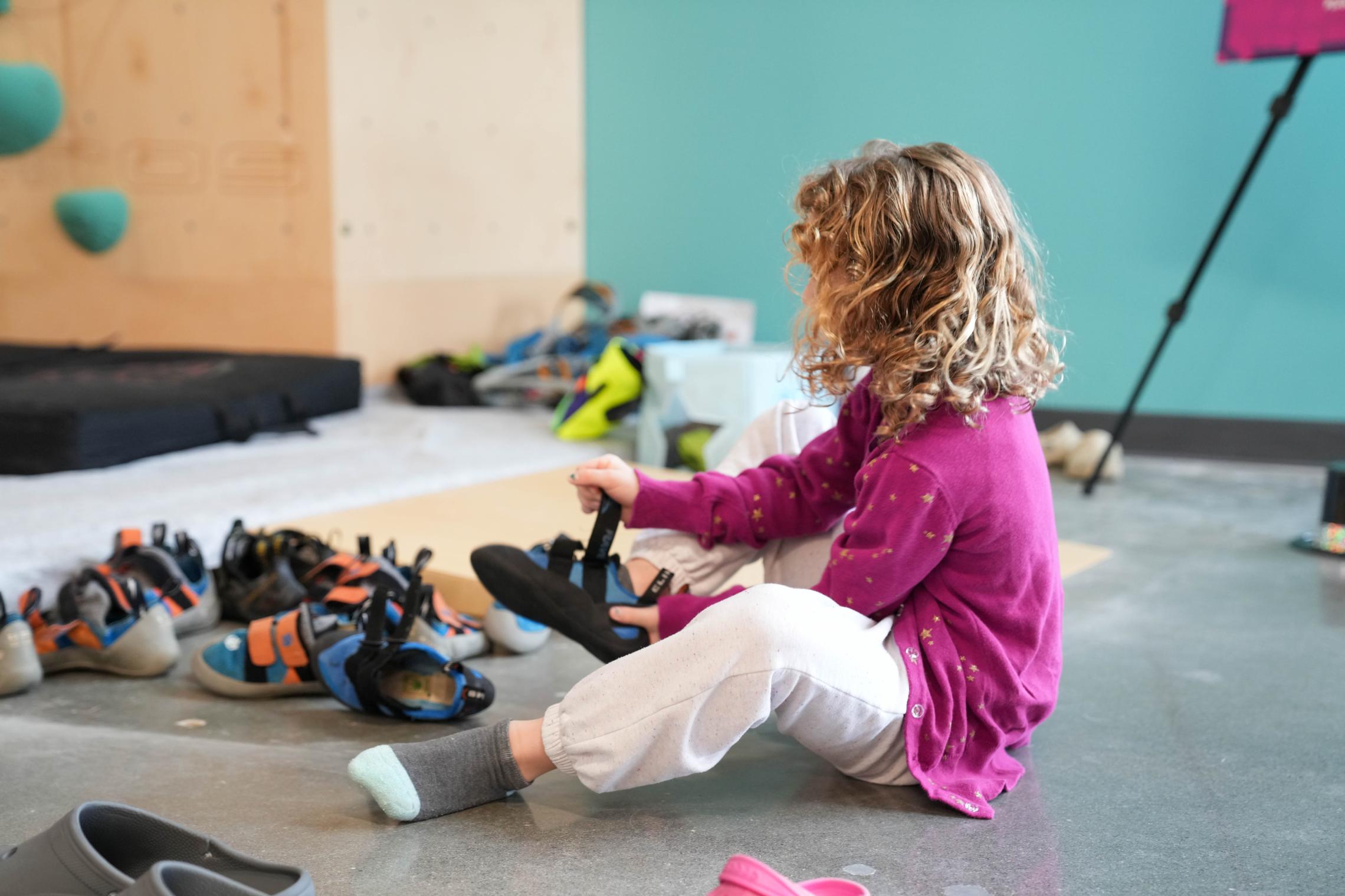 A child puts on climbing gear before using the Liz Rocks Climbing Wall at the Pearl Street Family YMCA, preparing for a youth adventure and confidence-building activity.