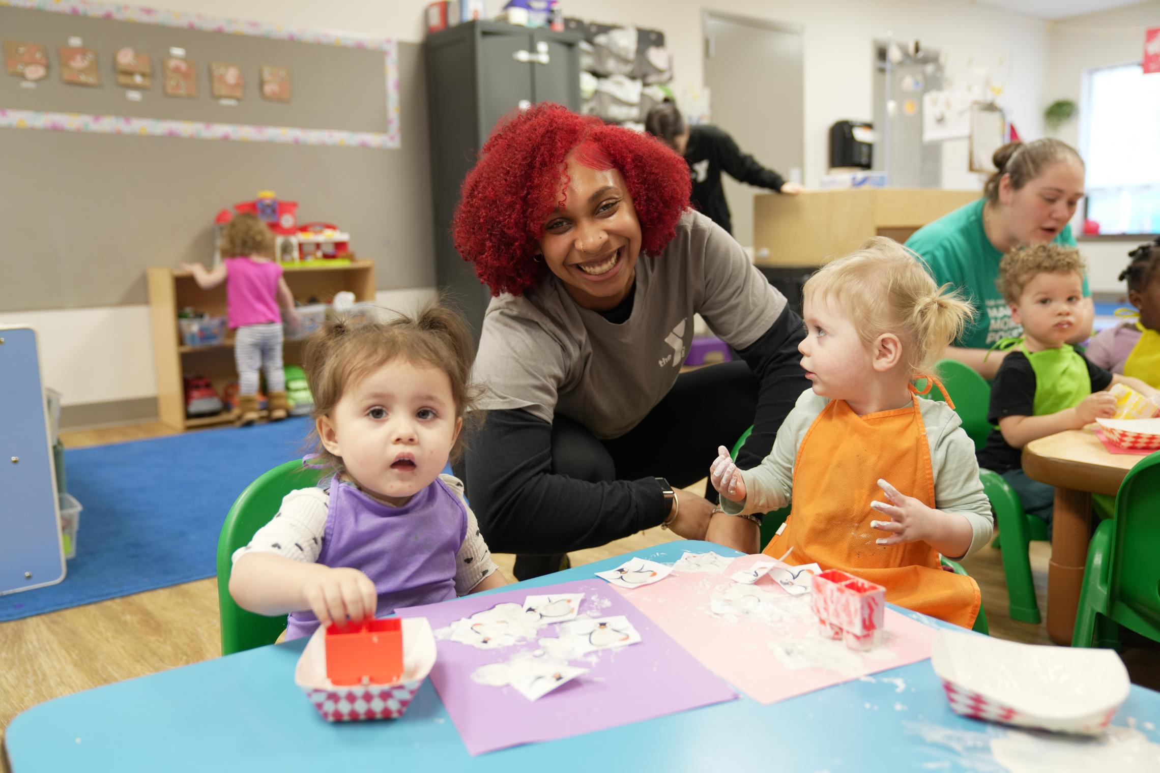 Children participate in an art activity at an early learning classroom table while an educator assists nearby. Craft materials, paper projects, and child-sized furniture are visible throughout the classroom, with additional children and educators engaged in activities in the background.