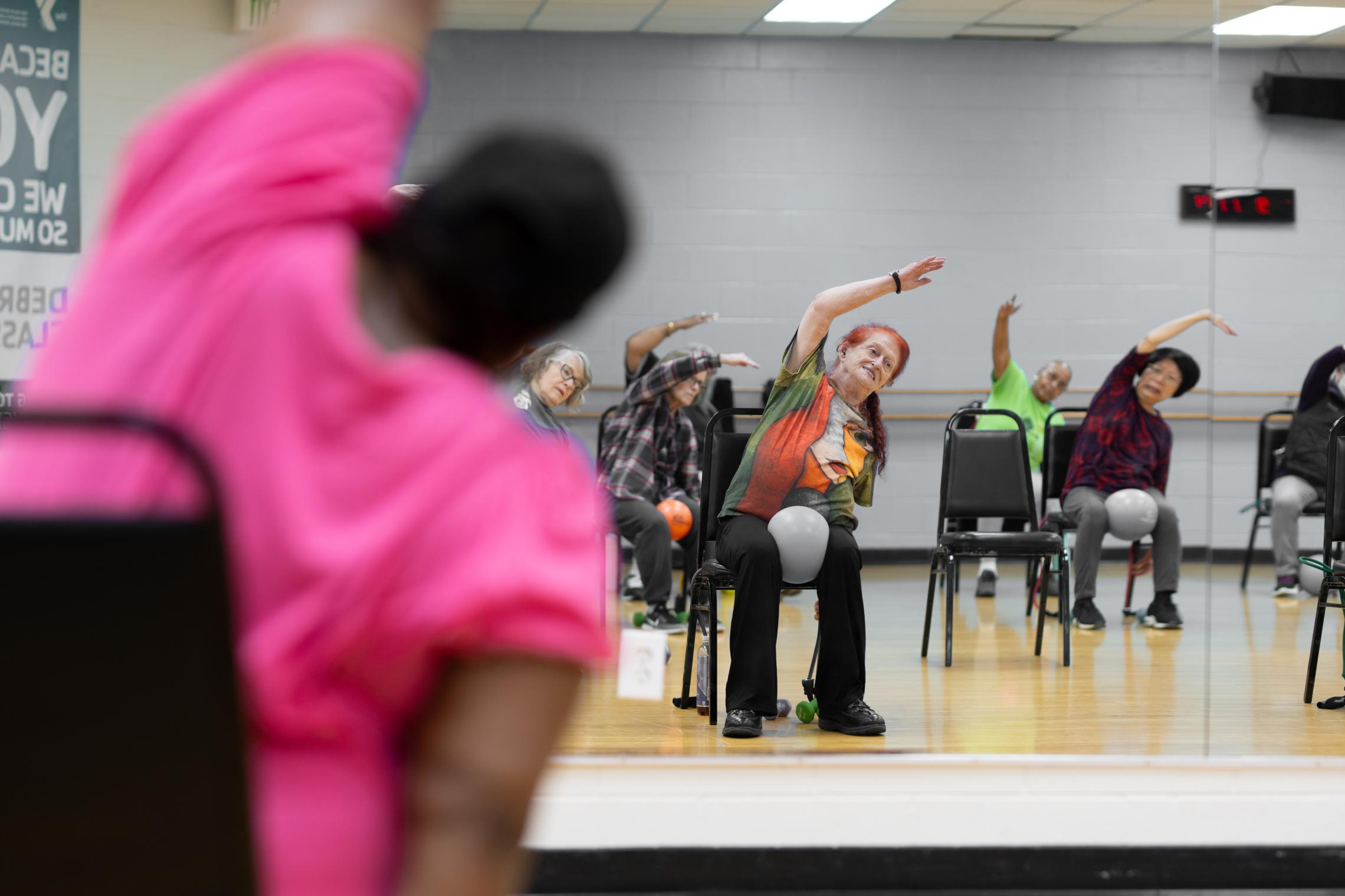 Participants in a YMCA chair-based fitness class perform seated side stretches while seated in chairs arranged across a studio. A wall mirror reflects the group exercising, with hand weights and exercise equipment visible around the room.